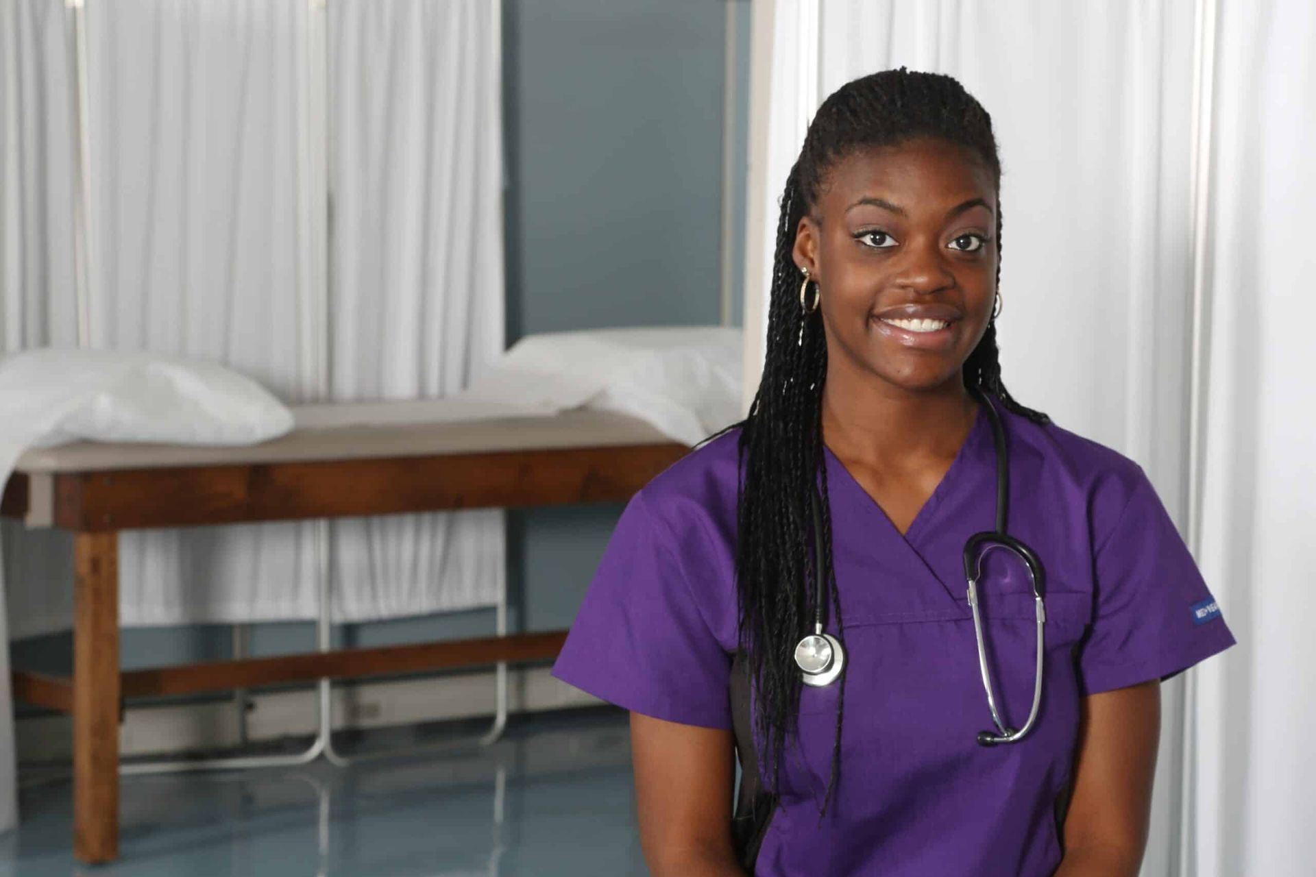 Nurse in purple scrubs smiling in a medical exam room.