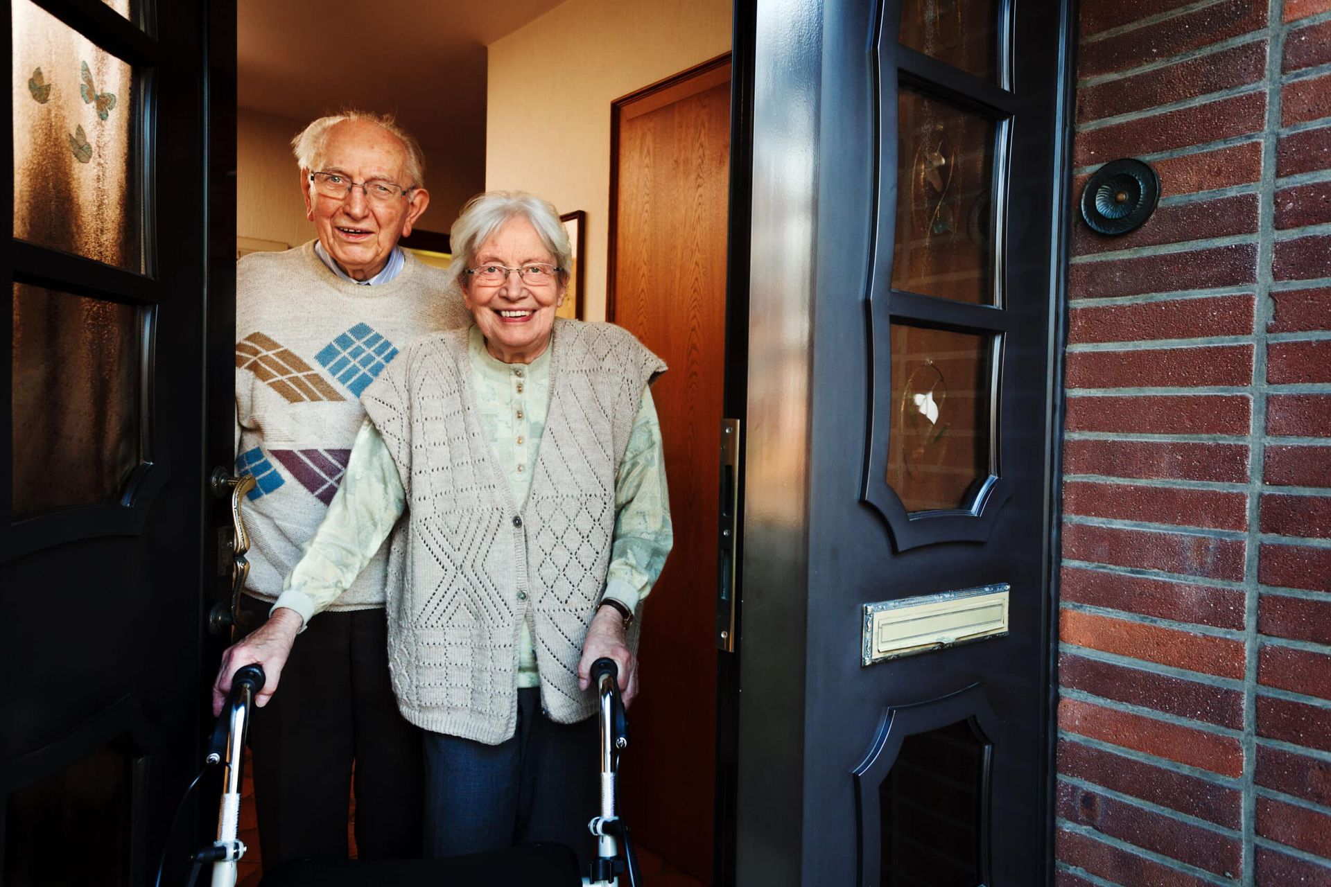 Elderly couple smiling at doorway, man in sweater, woman with walker. Brick house exterior.
