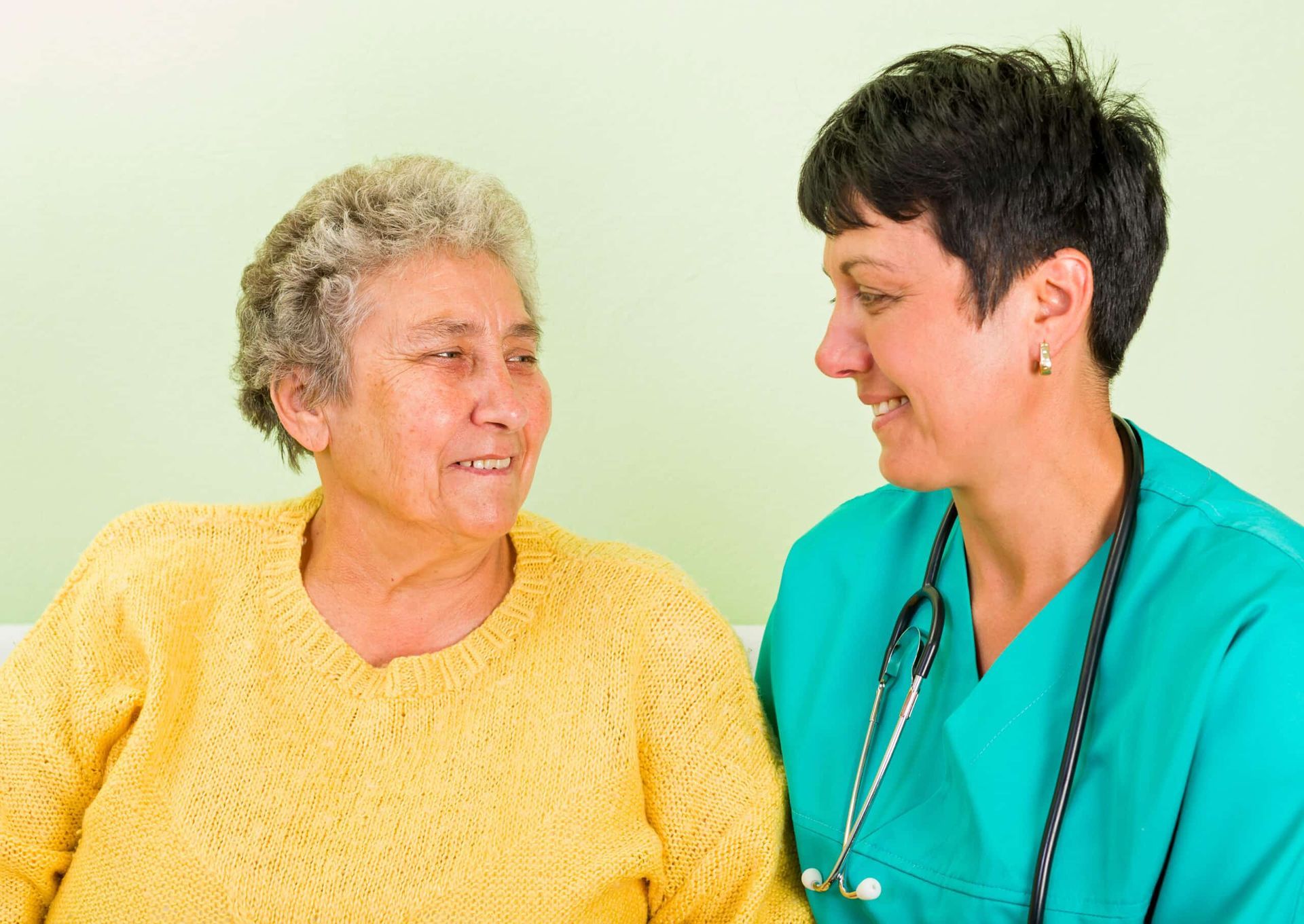 A smiling nurse in scrubs looks at an elderly woman wearing a yellow sweater, light green background.