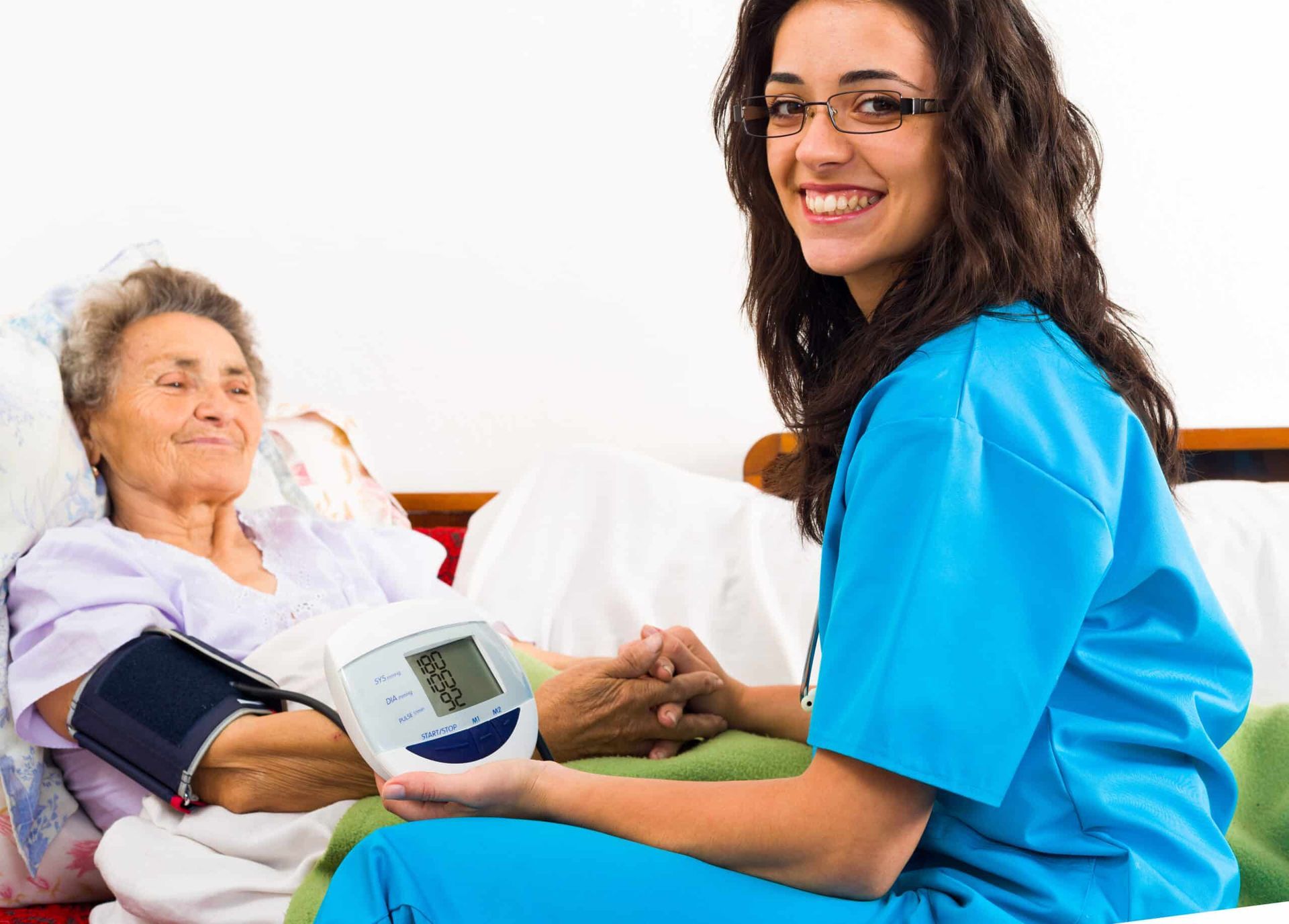 Nurse taking an elderly woman's blood pressure. Both are smiling. In a bed.