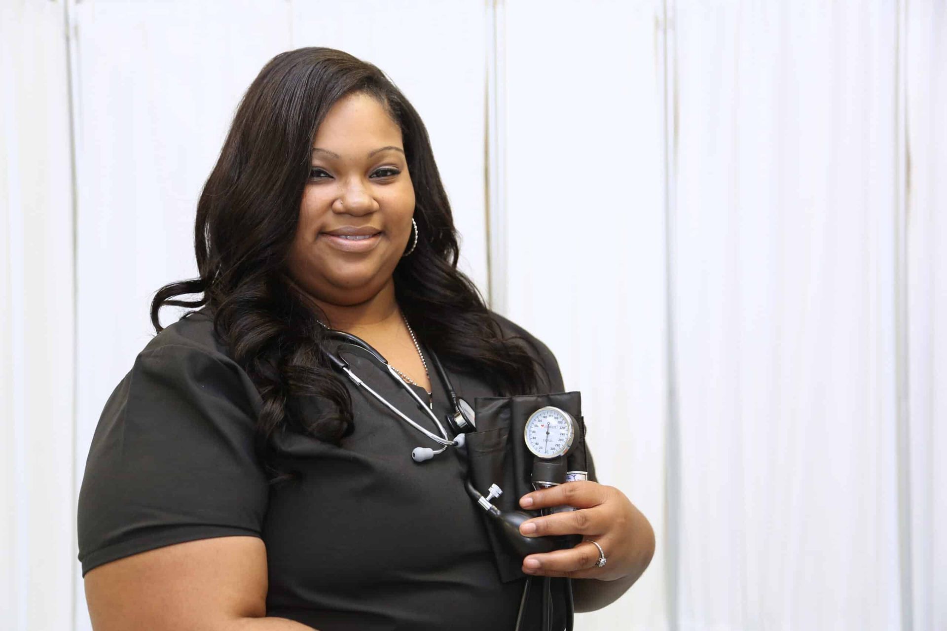Woman in black scrubs holds a blood pressure cuff and stethoscope, smiling. White curtain backdrop.
