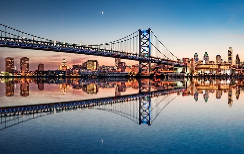 Philadelphia skyline at dusk, with the Ben Franklin Bridge and its reflection in the calm water.