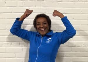 Woman in blue jacket with arms raised in excitement against a white brick wall.