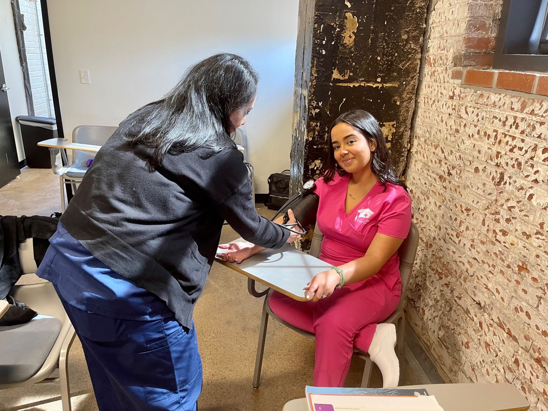 A woman in a pink scrubs has her blood pressure taken by another woman. Inside, office setting.