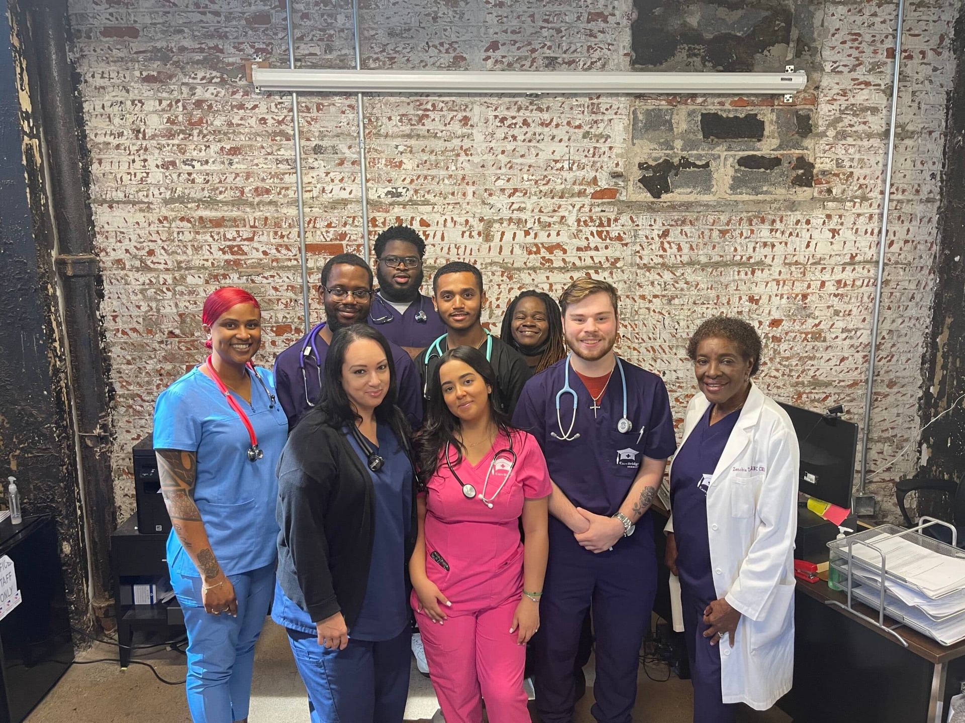 Group of medical professionals smiling, posing together in a room with a brick wall and some office equipment.