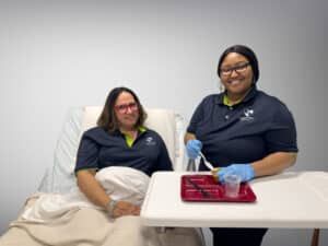Woman in bed, another assisting with food tray. Both smiling. In a hospital room setting.