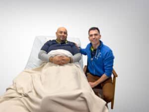 Man in bed with another man beside him. Both are smiling.
