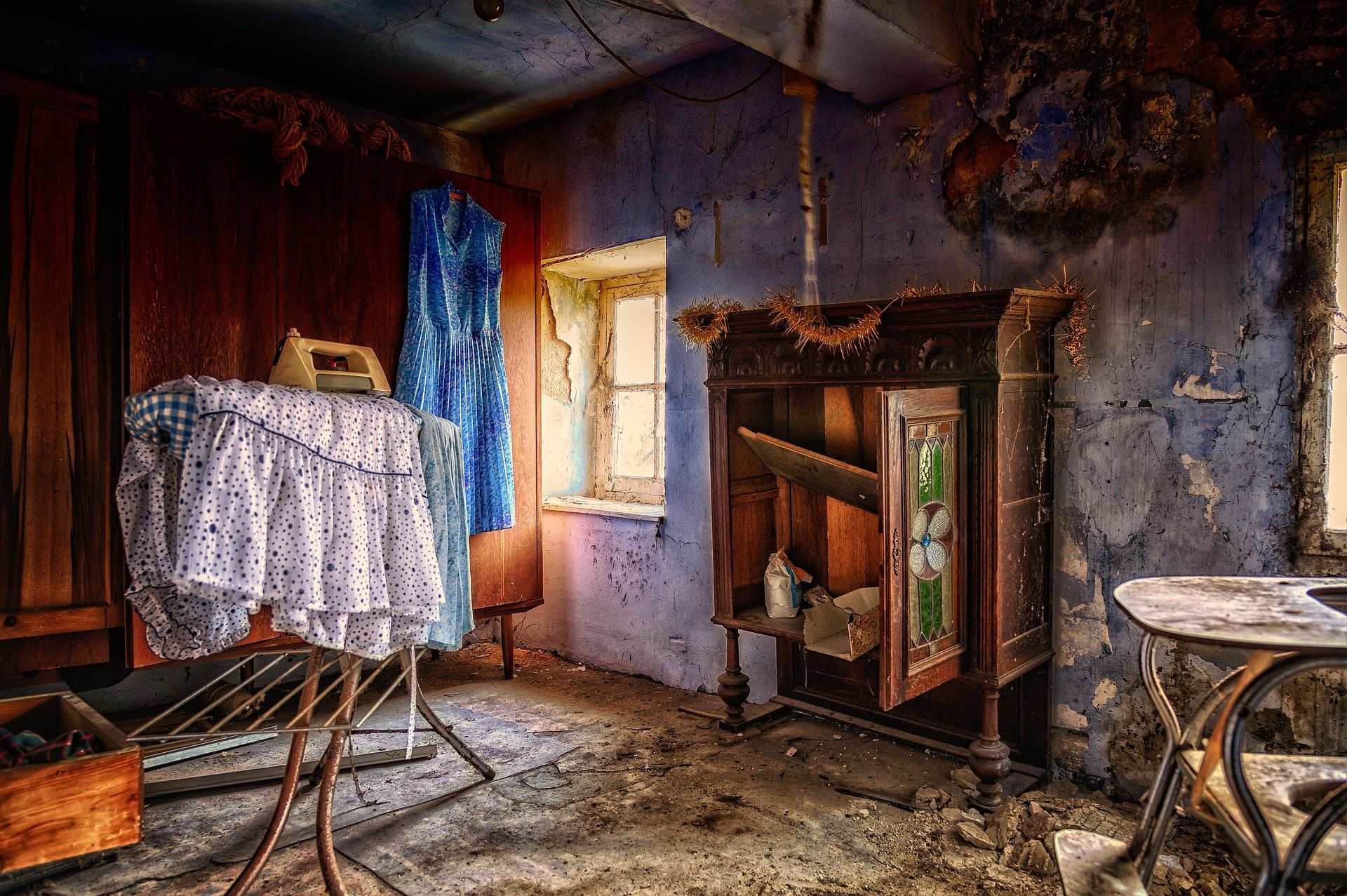 Abandoned room with peeling blue walls, weathered furniture, and a dusty ironing board.