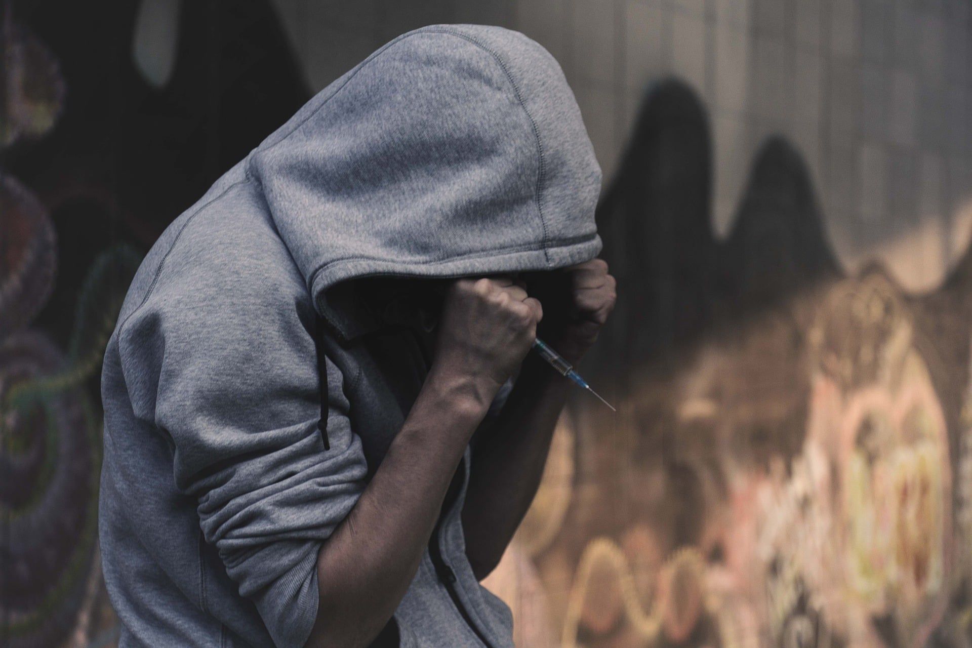 Person in a gray hoodie hunched over, face obscured, clenching fists. Graffiti-covered wall in the background.