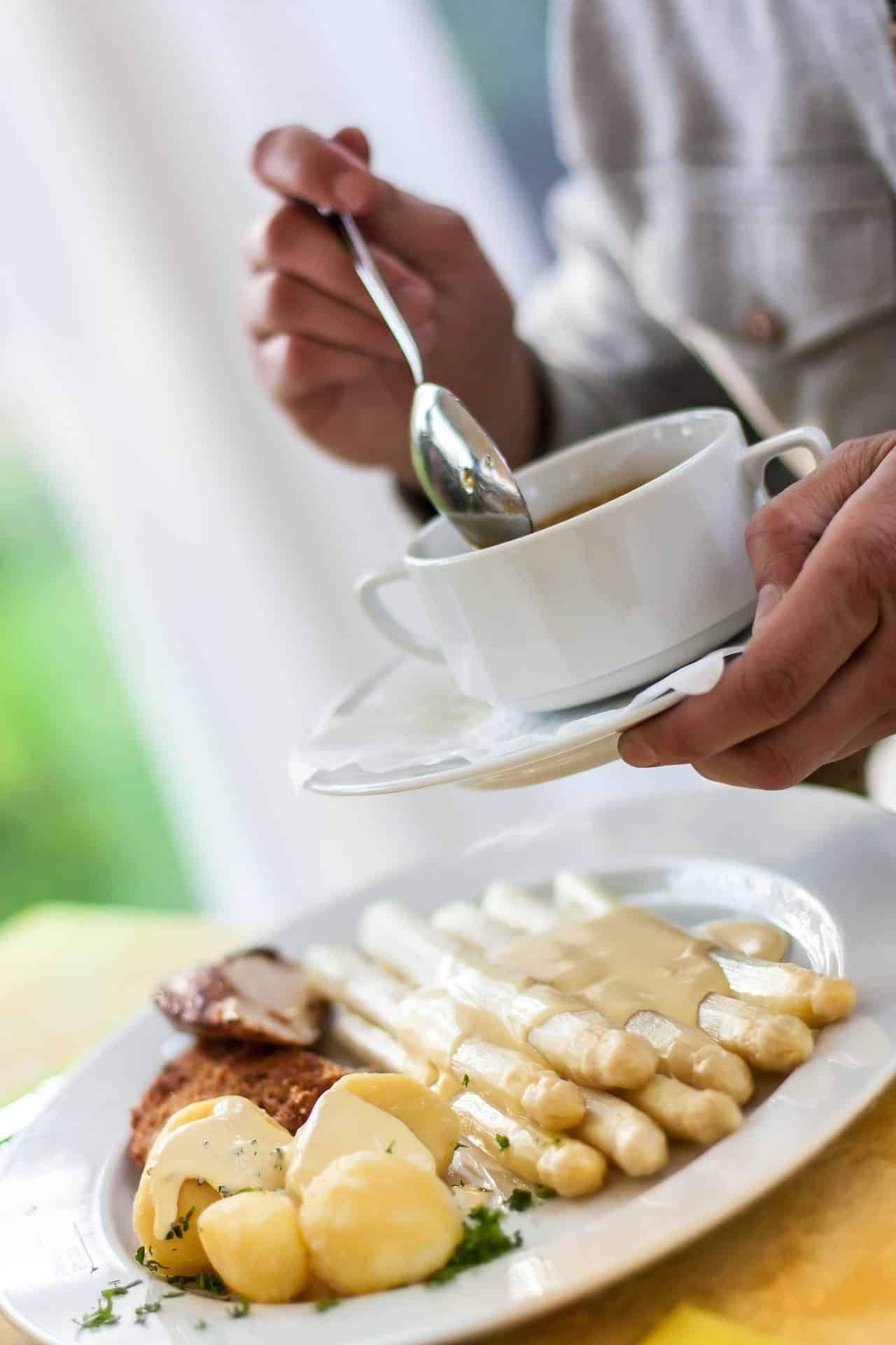 Person holding soup cup, stirrring. Plate of white asparagus, potatoes, and sauce.