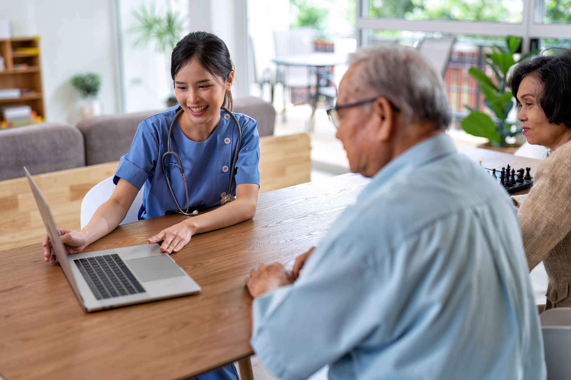Woman smiles while showing a laptop to two seated people at a table in a bright room.