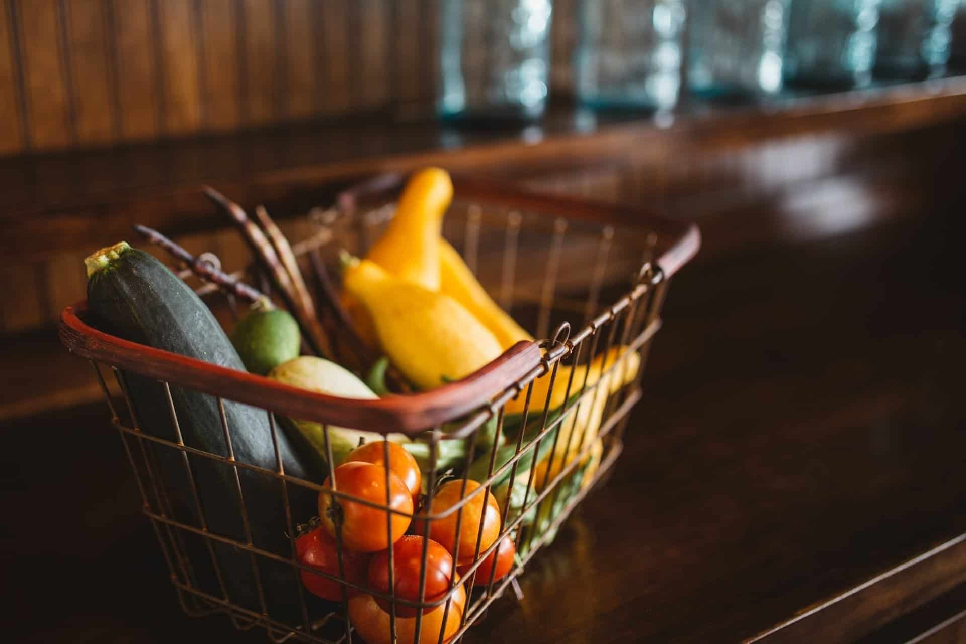 Vegetables in a metal basket on a wooden surface: tomatoes, zucchini, peppers, and more.