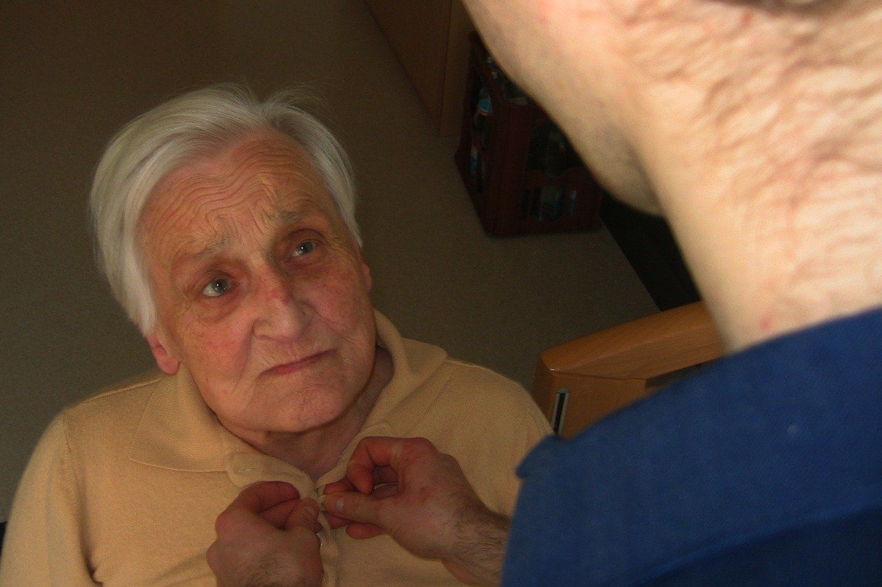 An elderly woman in a beige sweater has her buttoned by a person wearing blue, indoor setting.