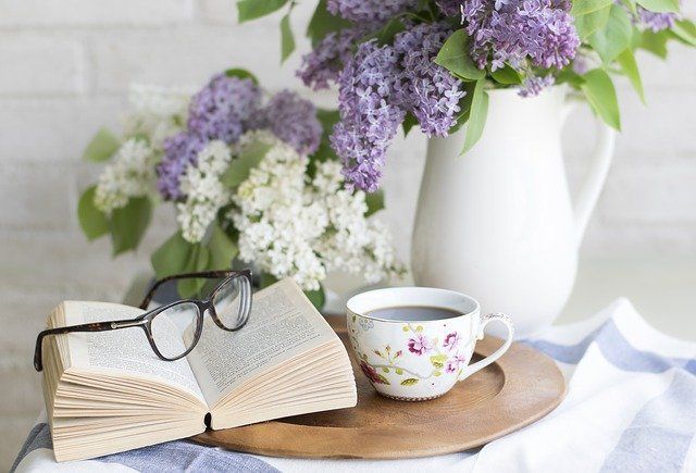 Open book with glasses, floral teacup, and lilac bouquet on a tray.