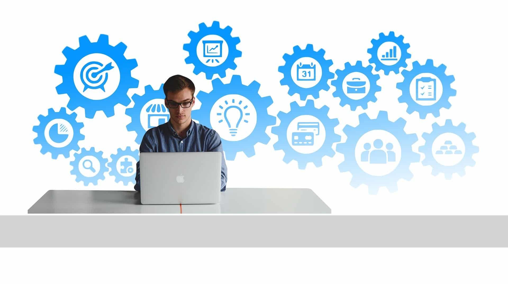 Man at laptop surrounded by blue gear icons representing processes, on white desk.