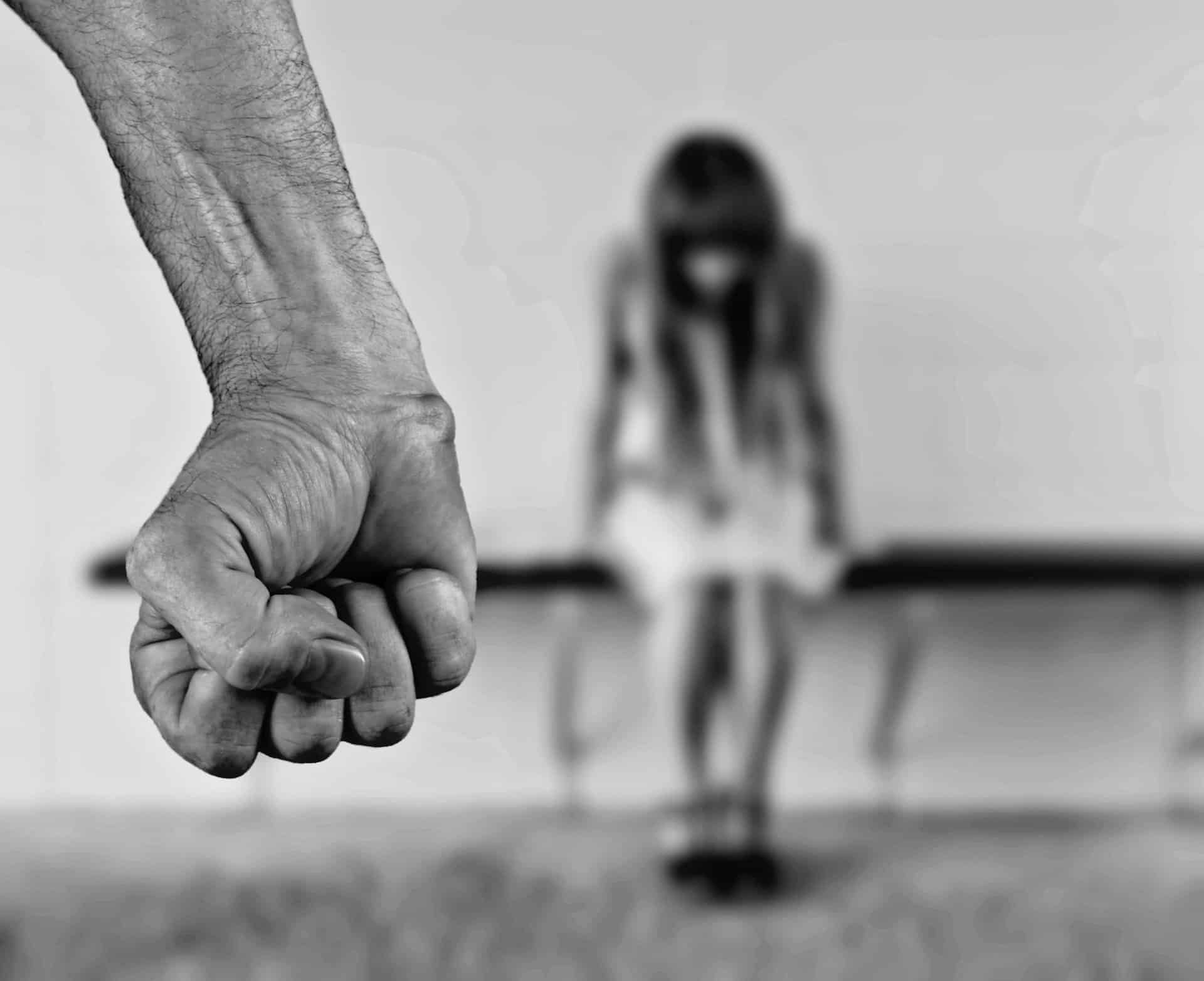 Fist clenched, raised menacingly toward a blurred, slumped figure of a person sitting outside.