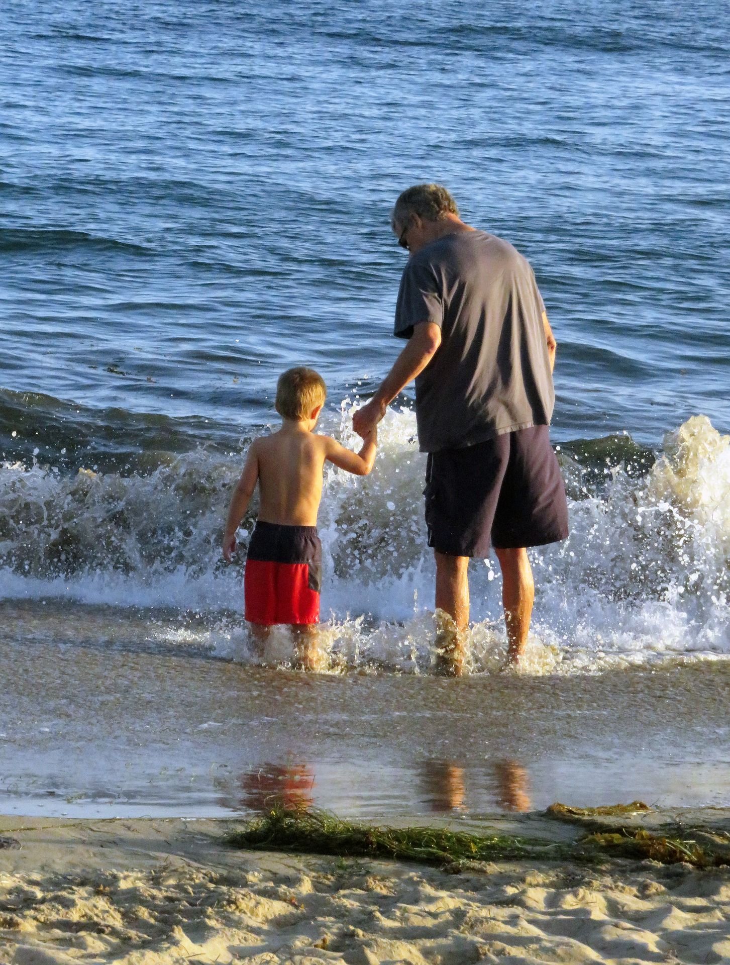 Grandfather holding hands with a young boy, both standing in the ocean waves at the beach.