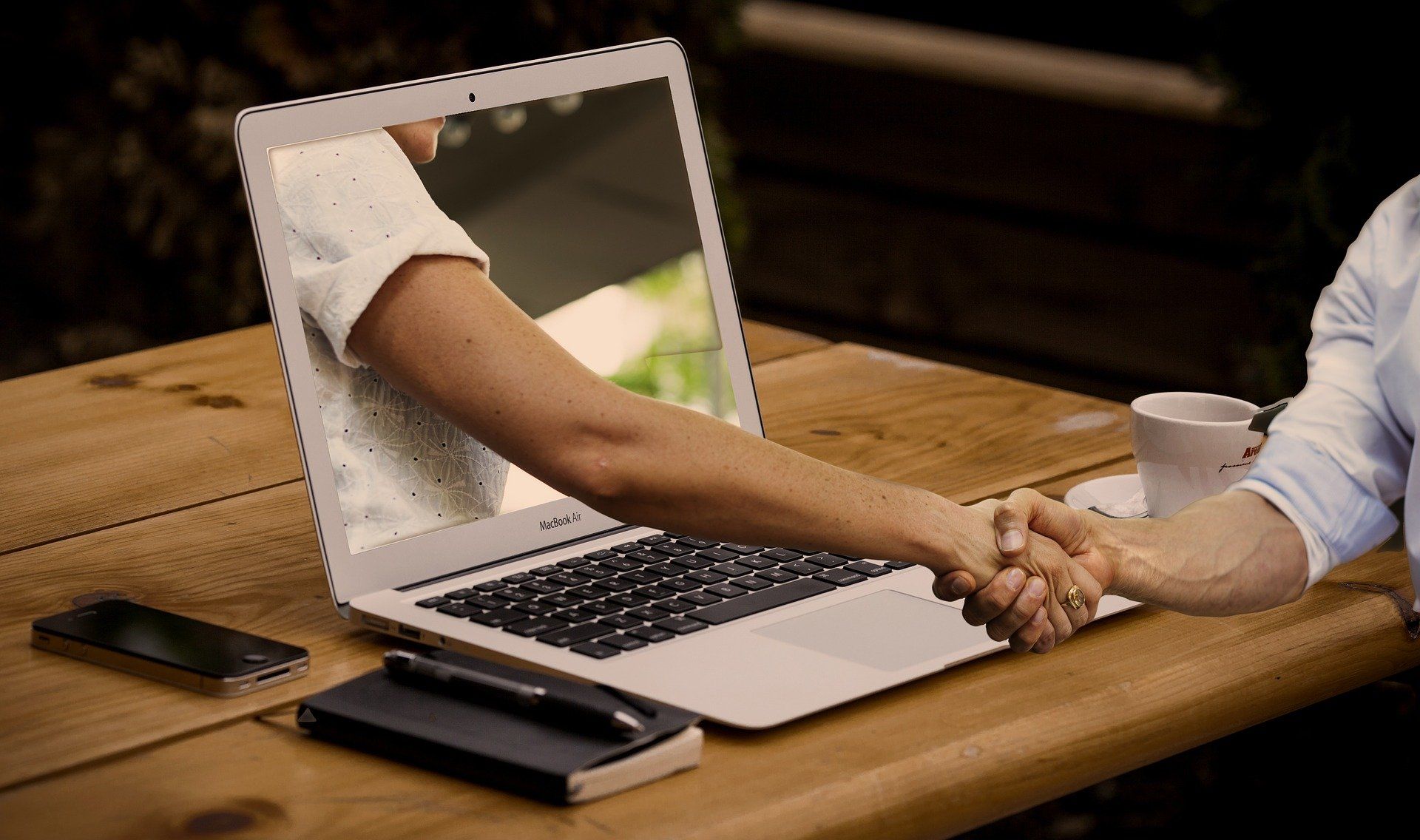 A hand reaches out of a laptop screen to shake hands with a person at a wooden table.