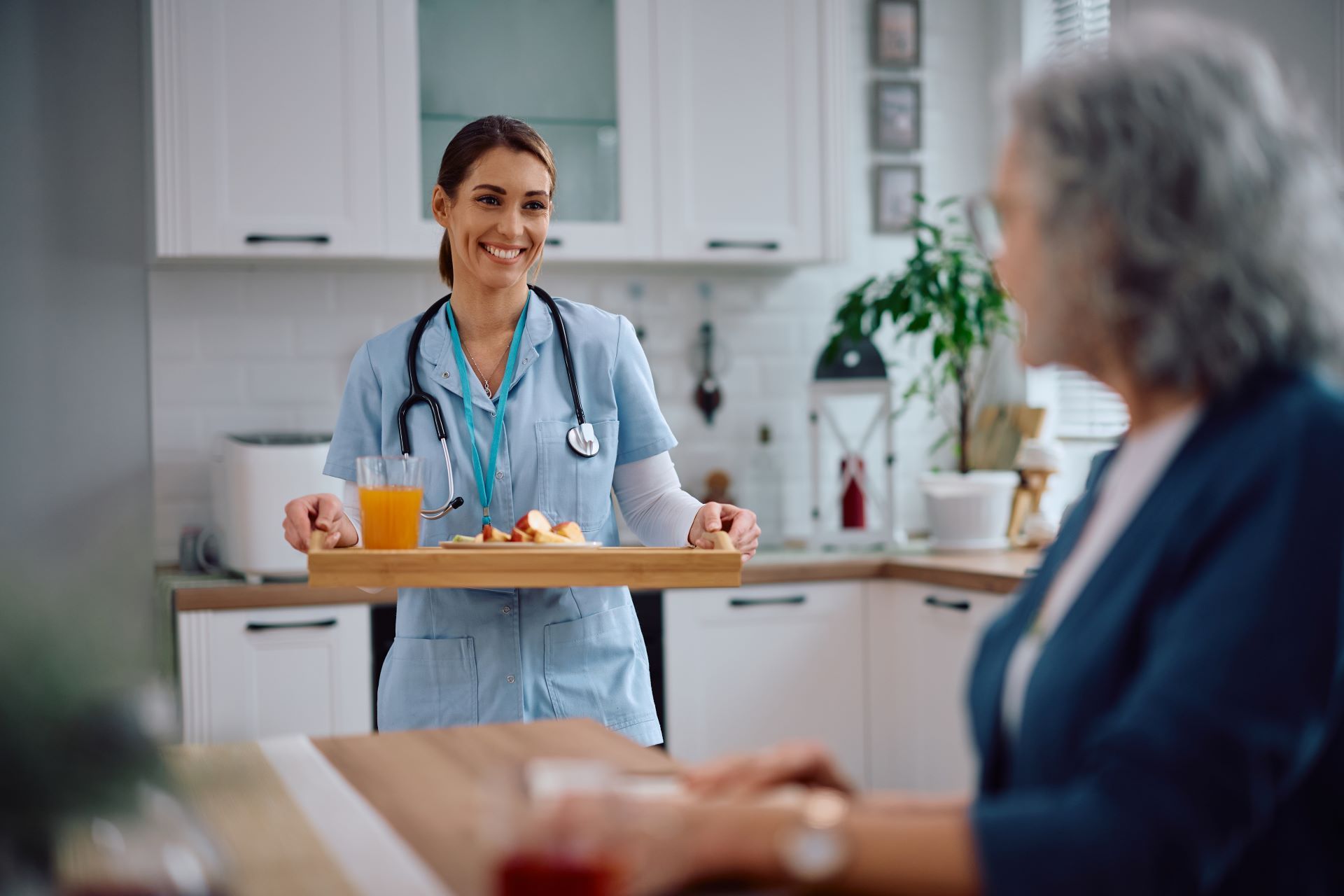 Caregiver smiles, serving breakfast to an older person at a kitchen table.