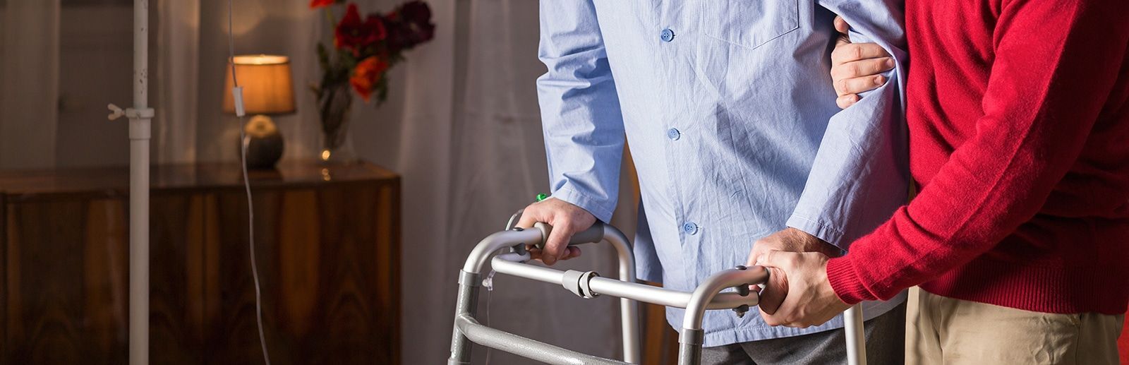Person using a walker being assisted by someone in a red sweater. Interior setting with a lamp and flowers.