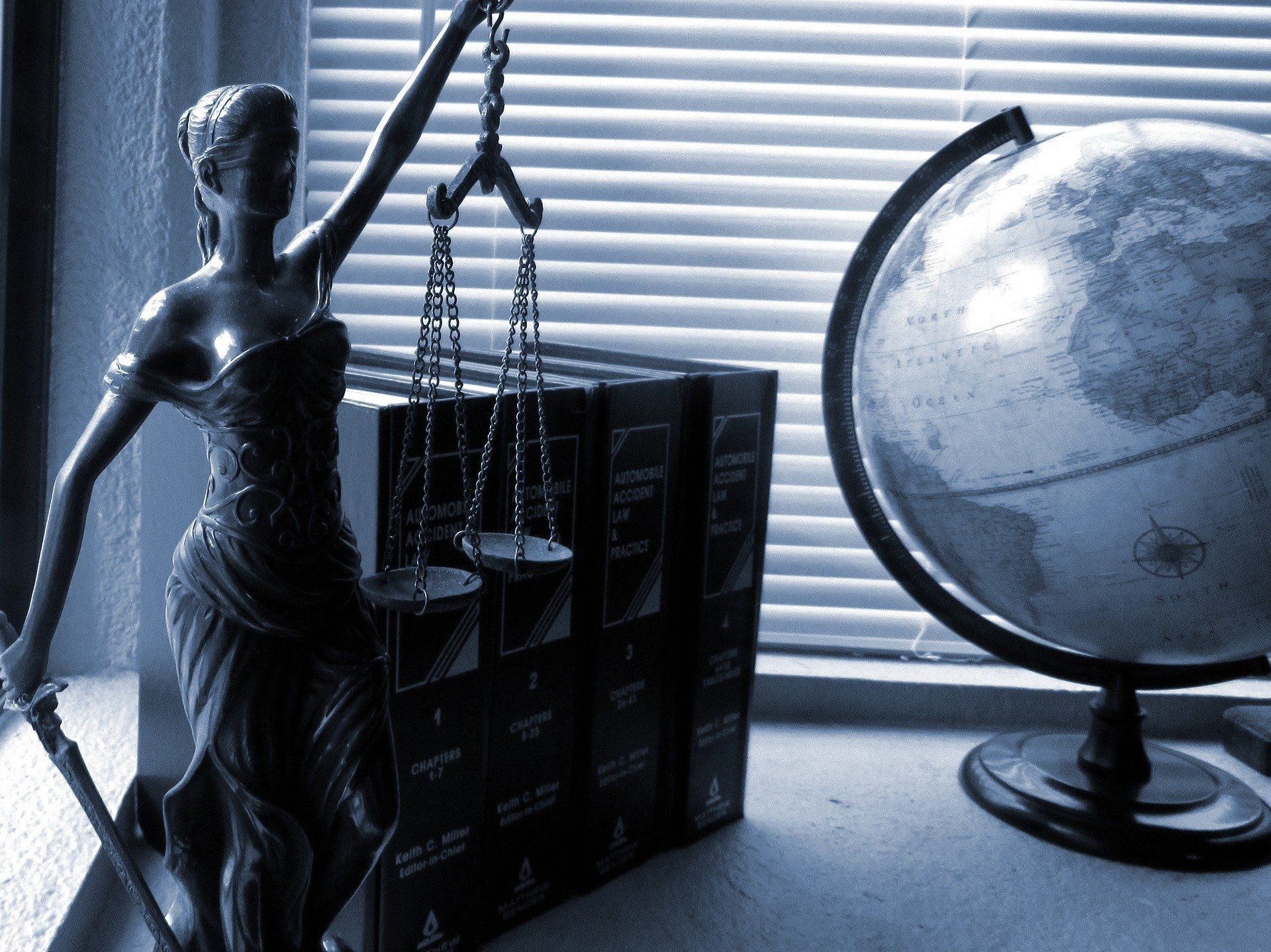Statue of Lady Justice next to law books and a globe, indoors near a window.