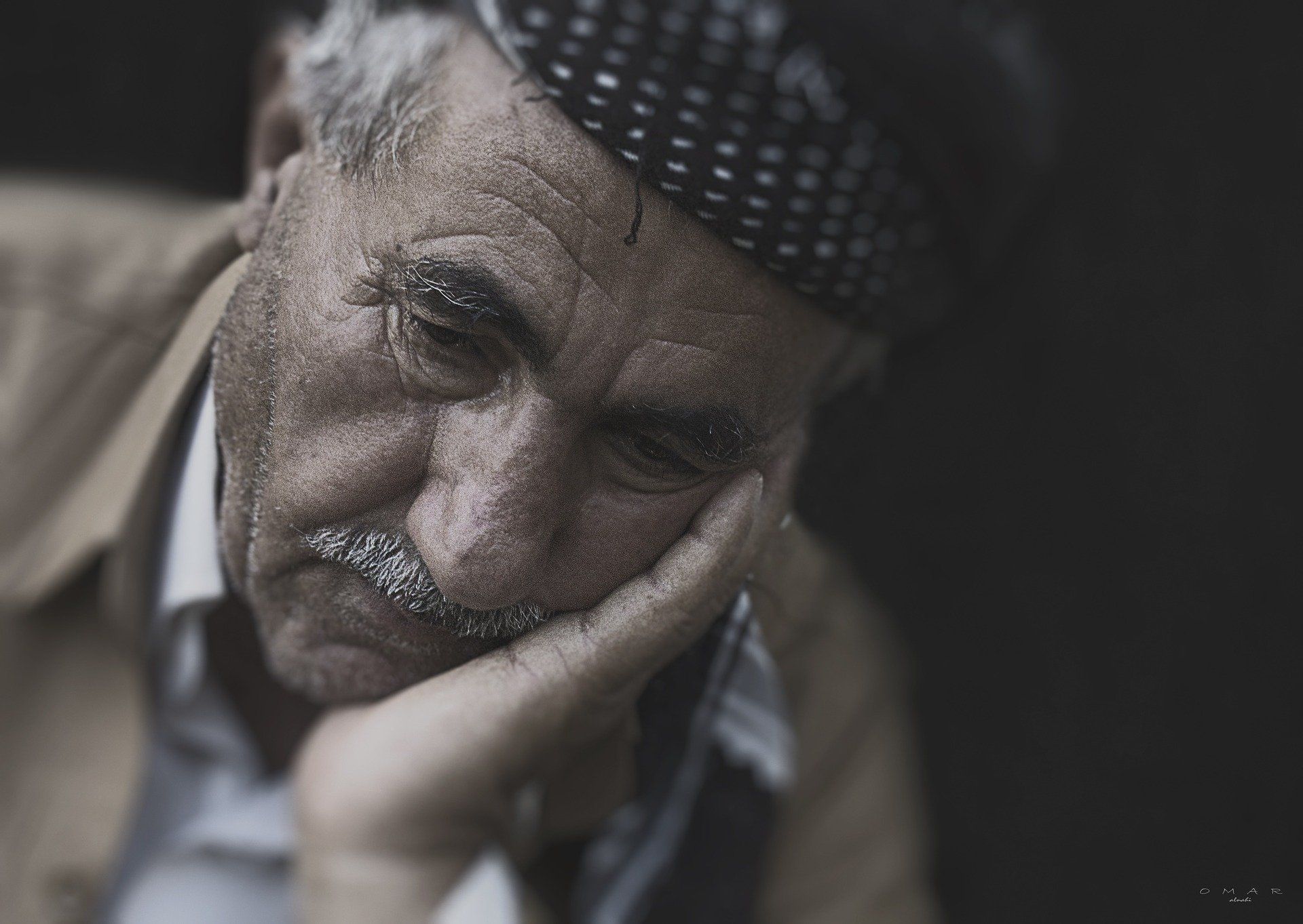 Elderly man resting his head on his hand, looking downcast. He has a mustache and a patterned cap.