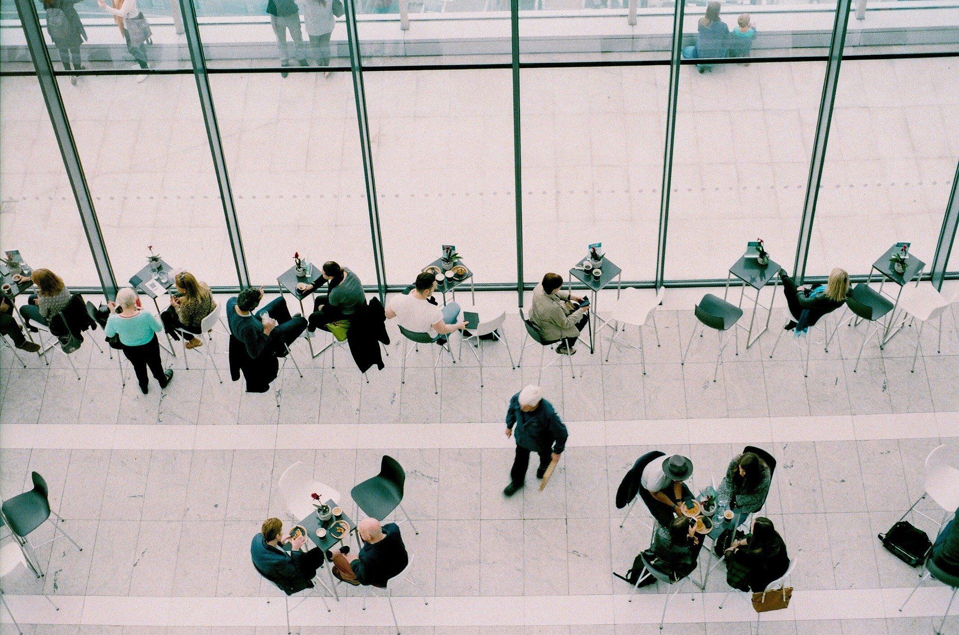 Overhead view of people at tables near large windows in a modern building.