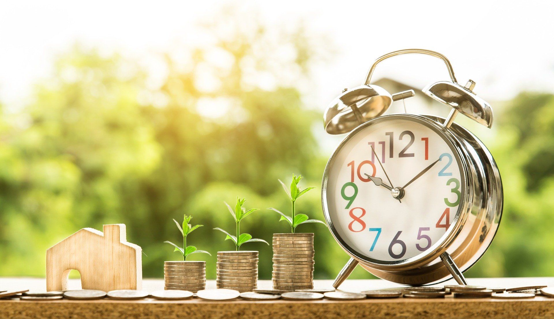 Wooden house and stacks of coins with growing plants, near a clock in a sunny outdoor setting, symbolizing financial growth.