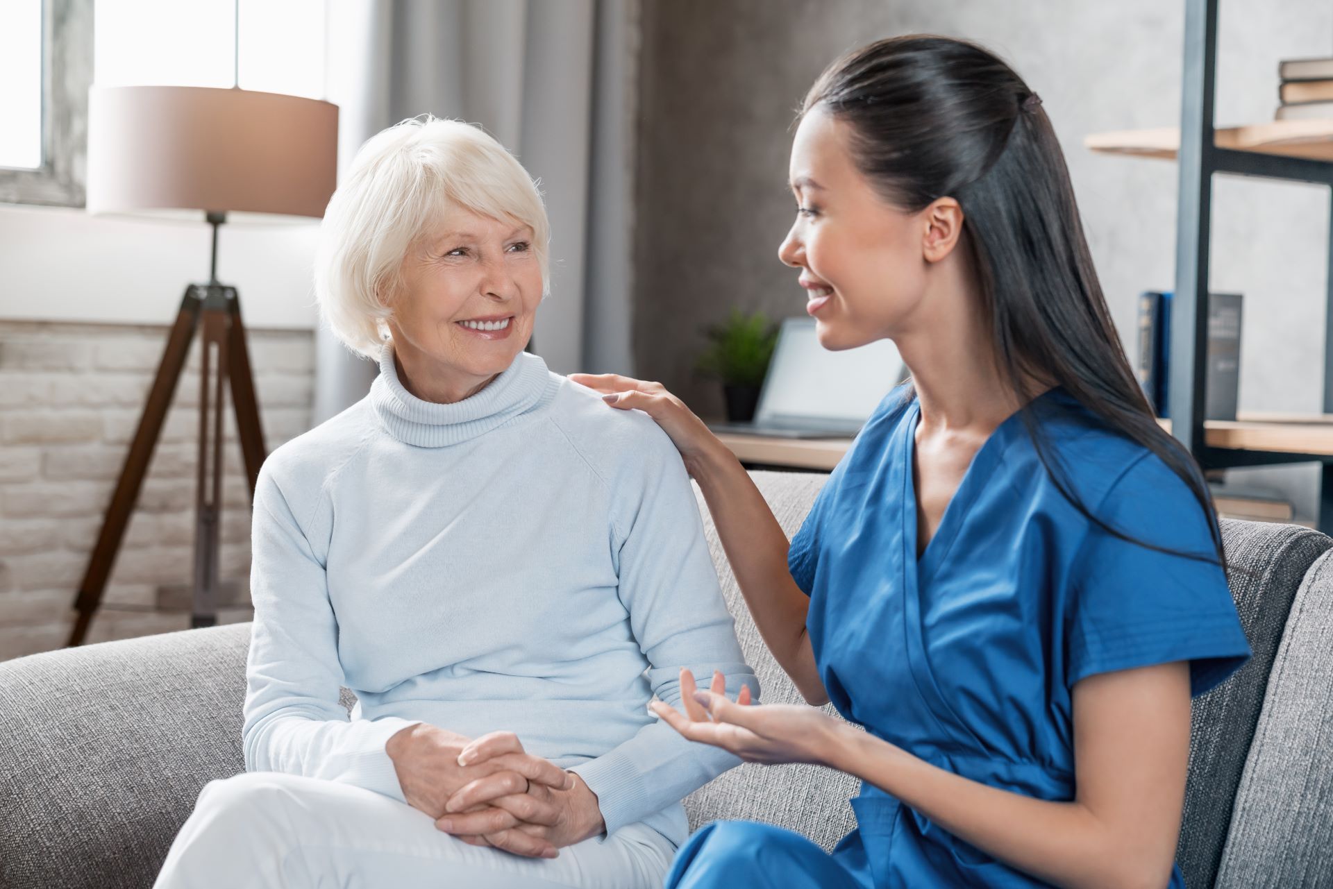 A caregiver in blue scrubs comforts an elderly woman on a sofa indoors, hand on her shoulder.