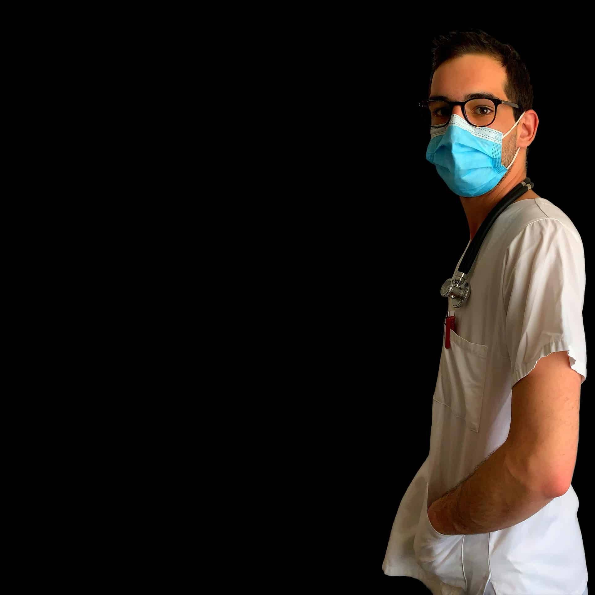 Man in scrubs and a mask, wearing glasses, a stethoscope, and looking to the side, with hands in pocket.