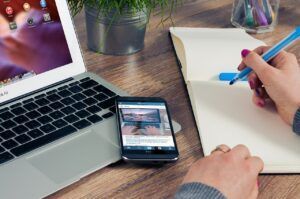 Person writing in notebook with pen next to laptop and smartphone on a wooden desk.