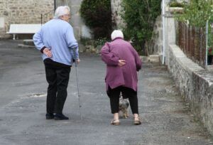 An elderly couple walking outdoors; the woman has a dog; both have their hands on their lower backs.