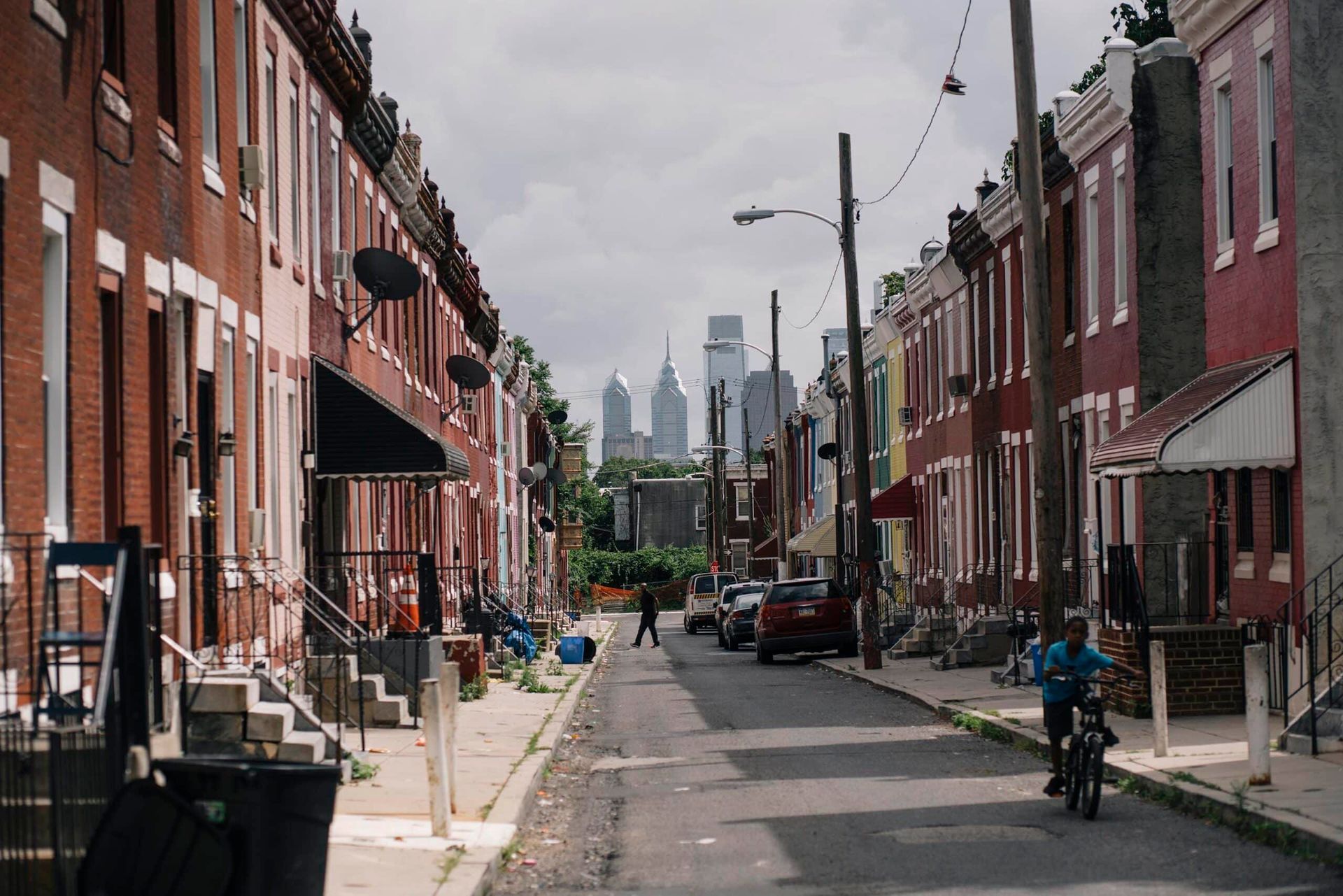 Rows of brick rowhouses line a city street, with the Philadelphia skyline visible in the distance.
