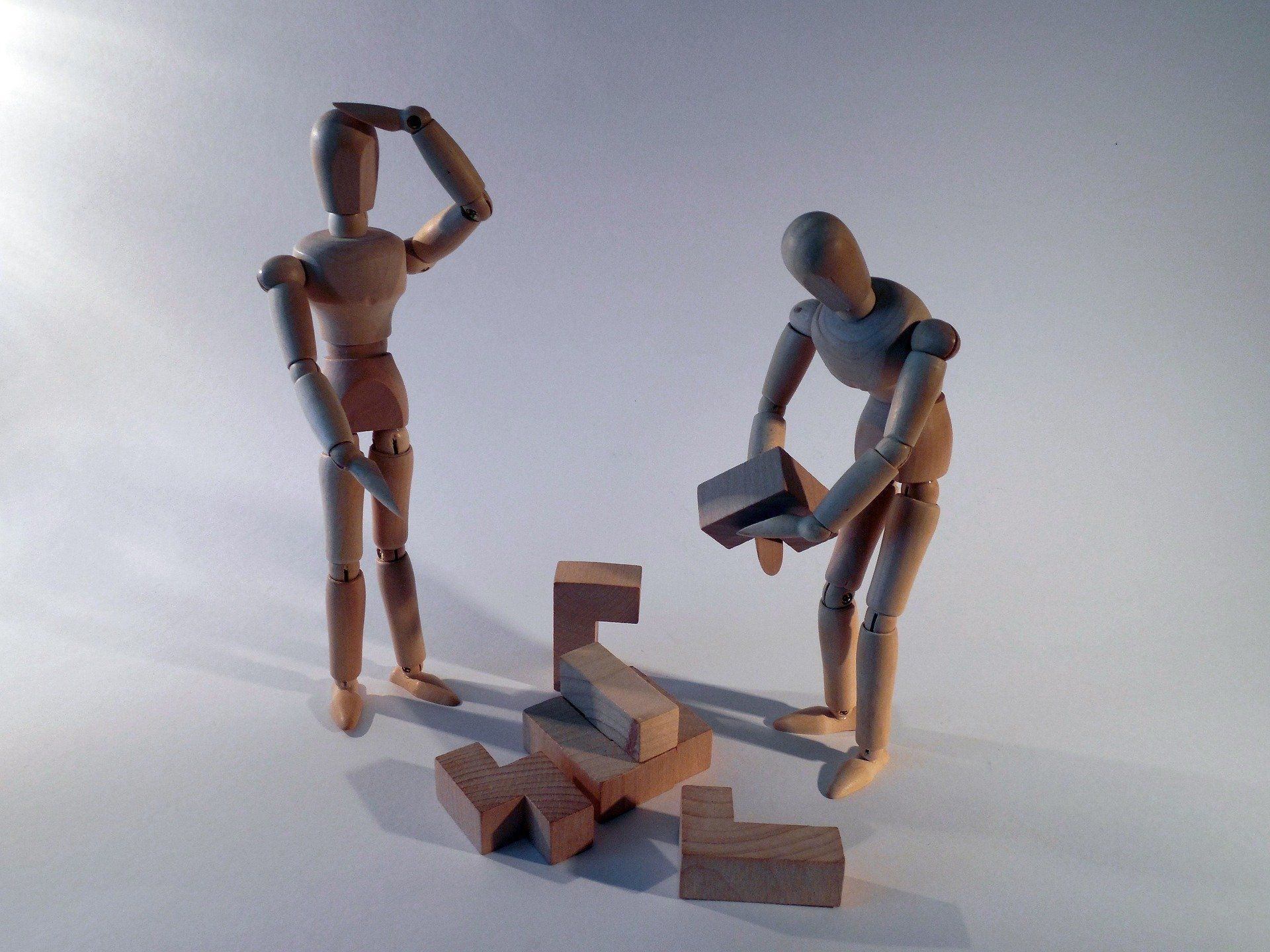 Two wooden mannequins building with blocks, one confused, the other focused. White background.