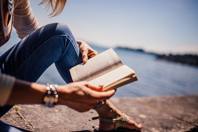 Woman reading a book outdoors by the water, in jeans and sandals on a sunny day.