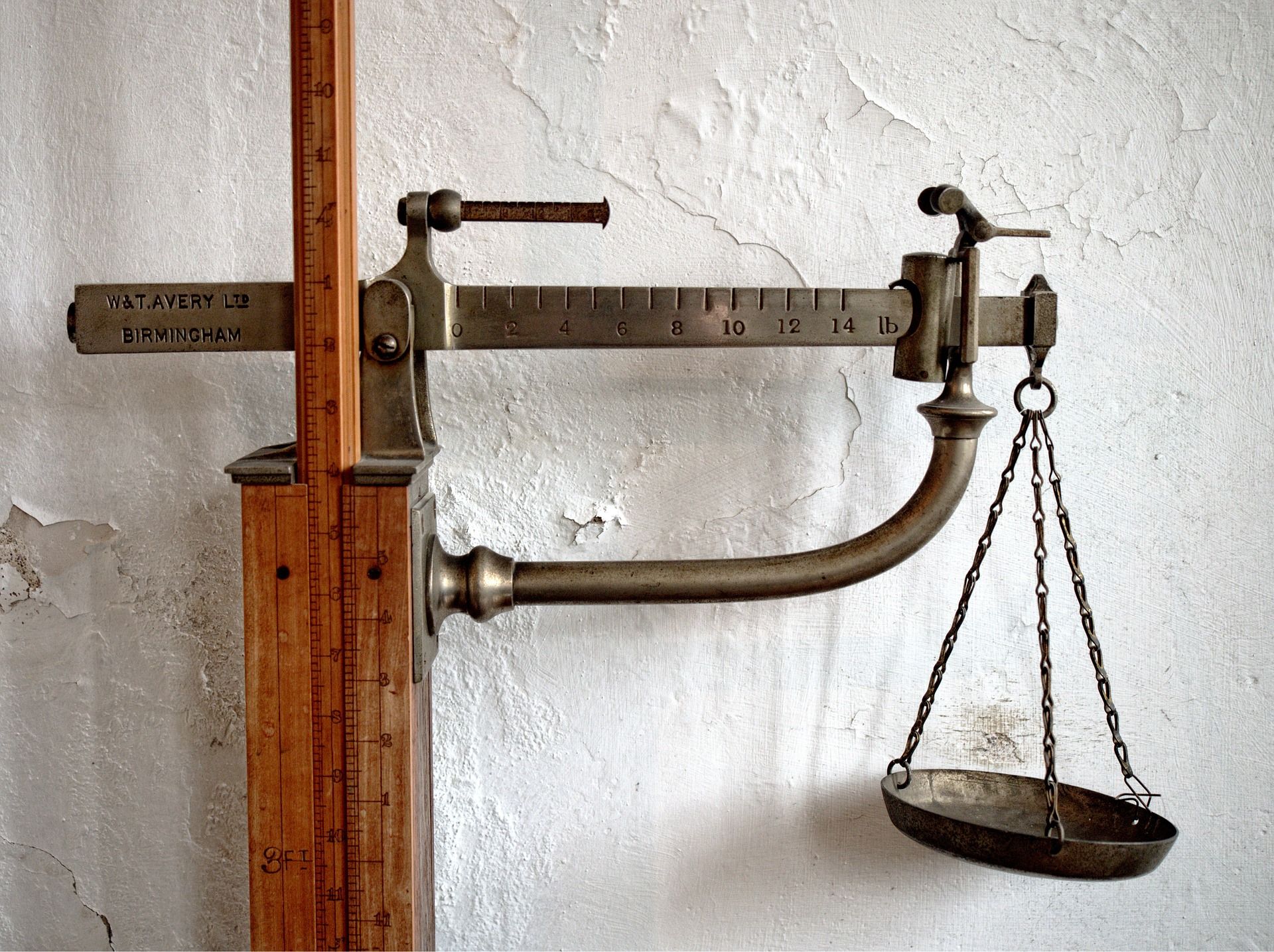 Antique scale with a bowl hanging from chains against a peeling, white wall.