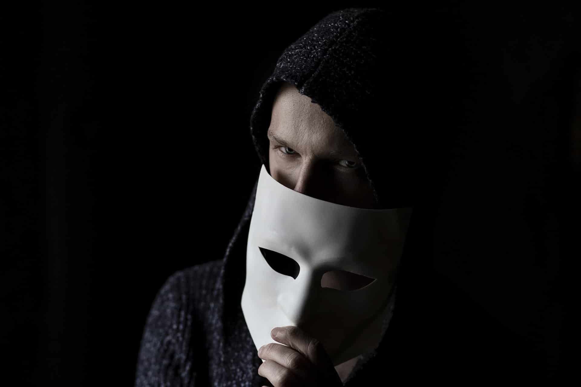 Person in dark hoodie holding a white mask, partially covering face.