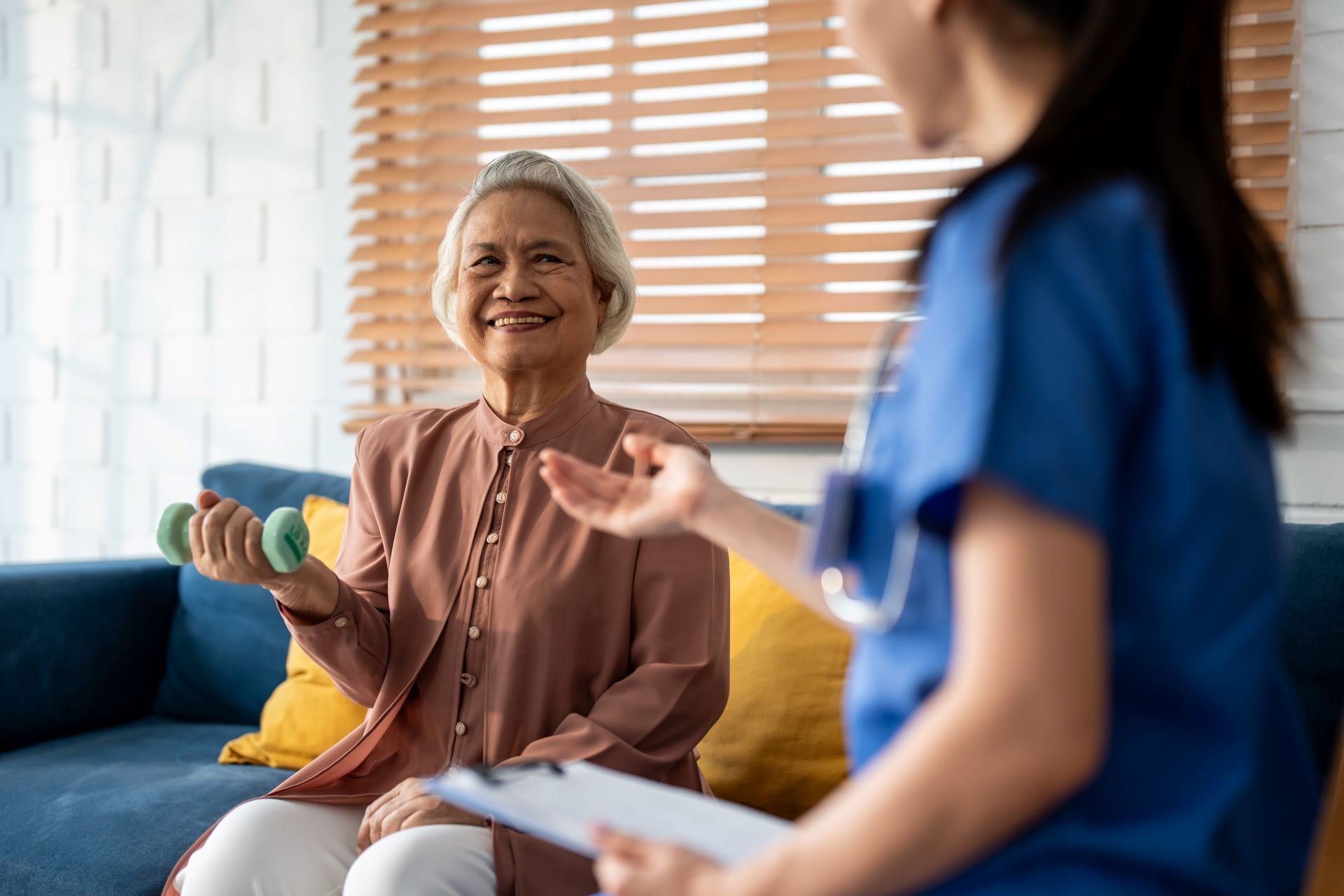 An older adult lifts weights, guided by a healthcare professional holding a clipboard in a living room.