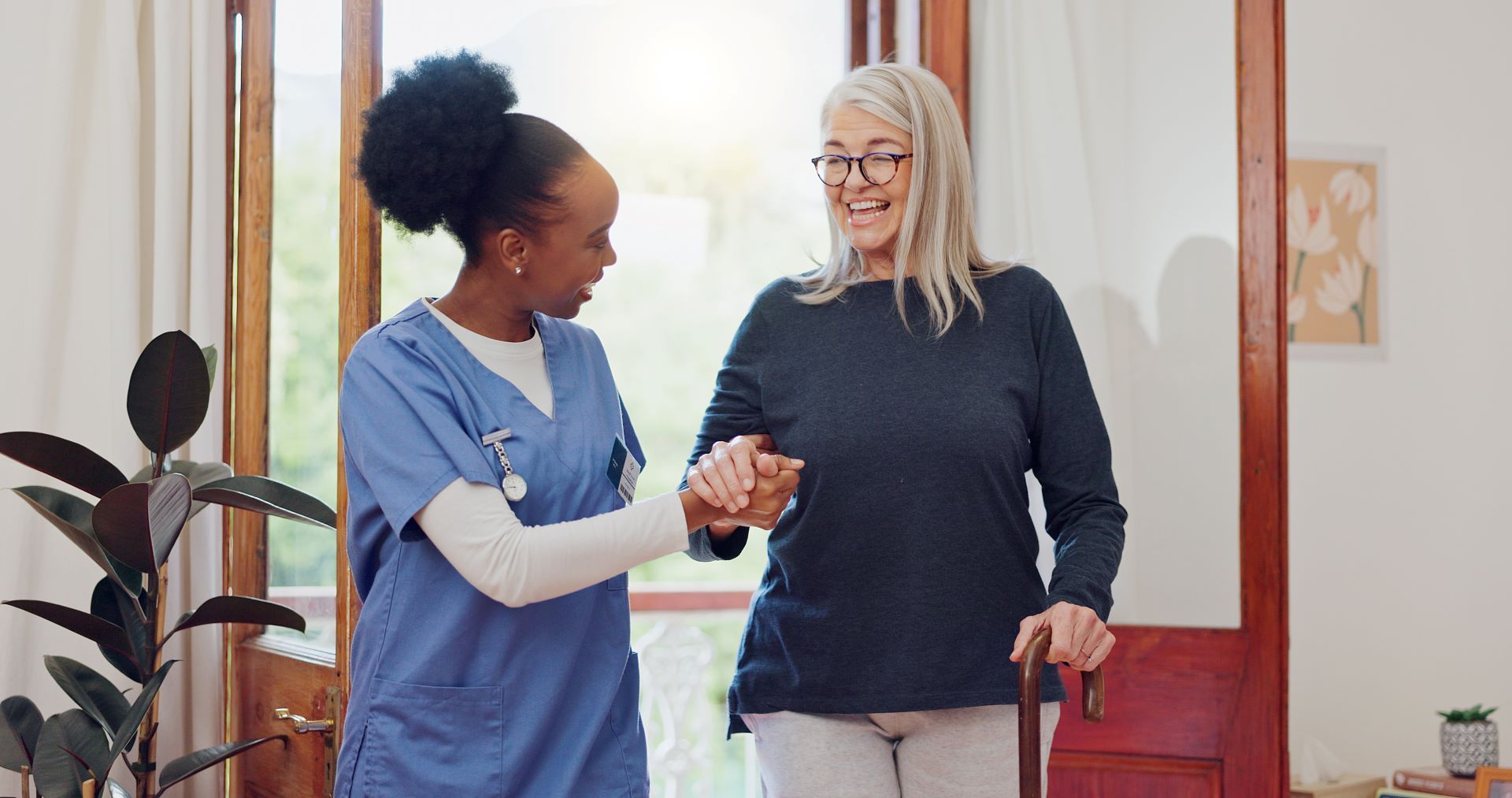 A healthcare worker in blue scrubs helps a smiling person walk indoors, holding their hand for support.
