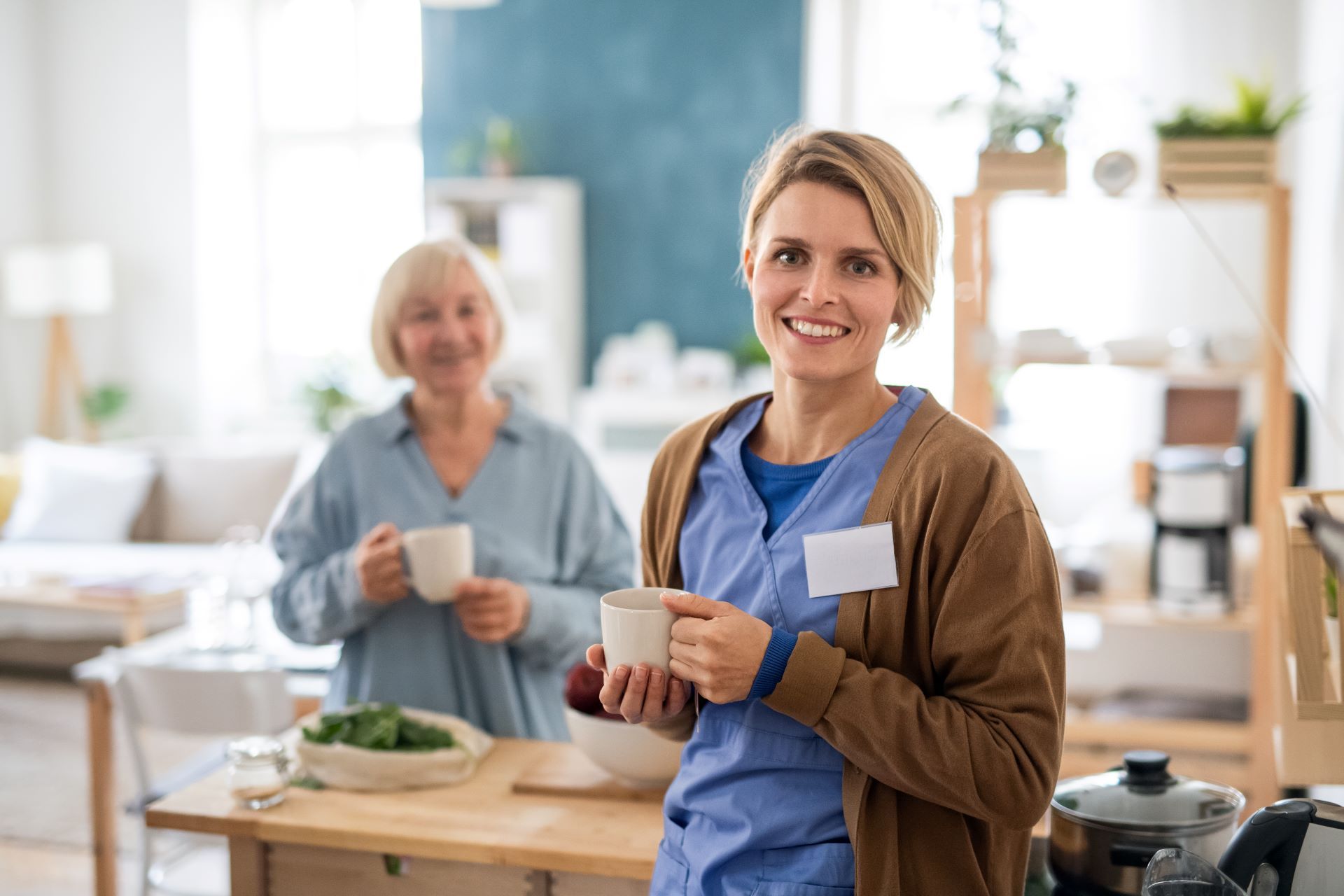 Woman in scrubs smiles, holding a mug, with another woman in the background. Kitchen setting.