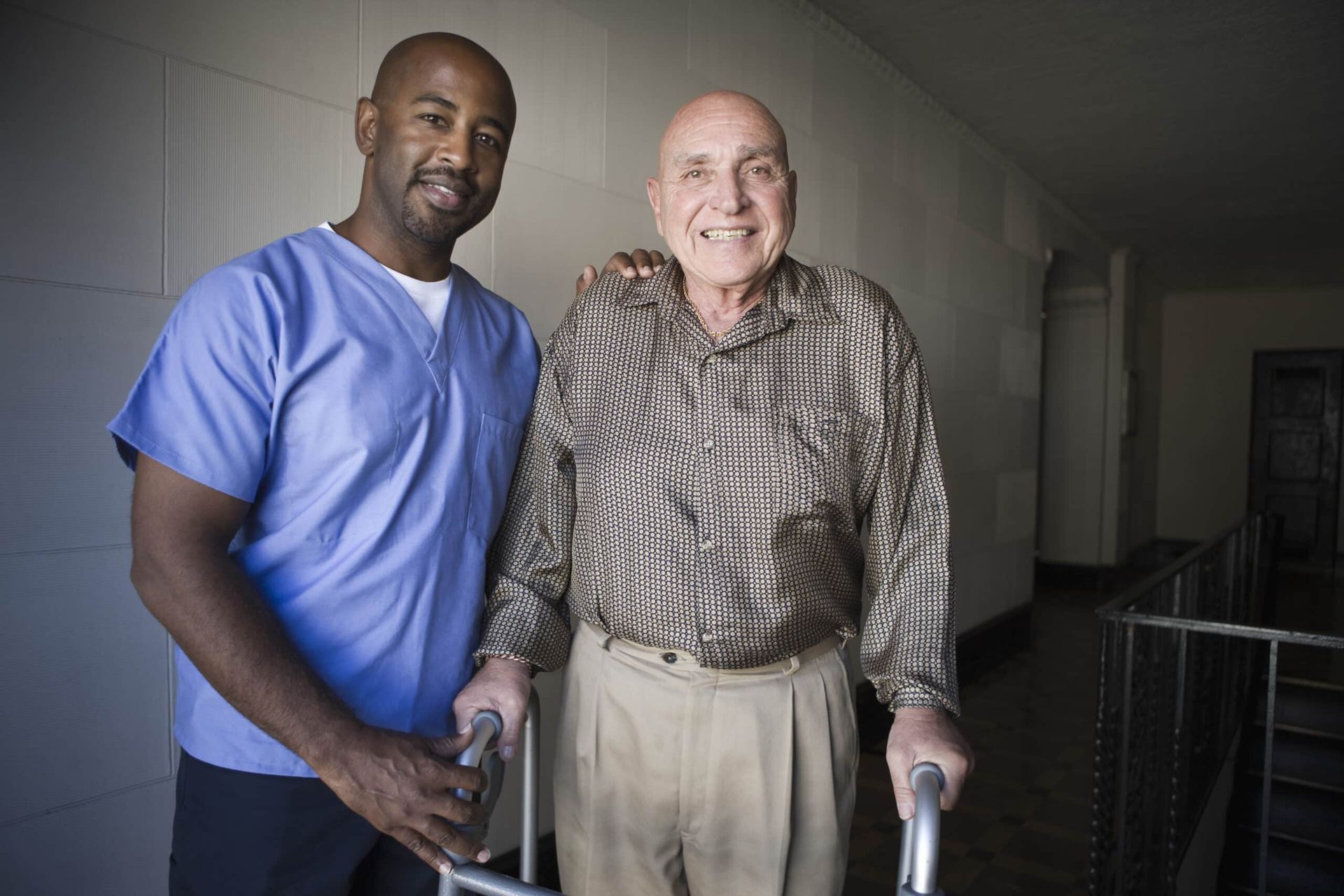 Caregiver assists an elderly man with a walker in a hallway. Both smile, and caregiver wears blue scrubs.