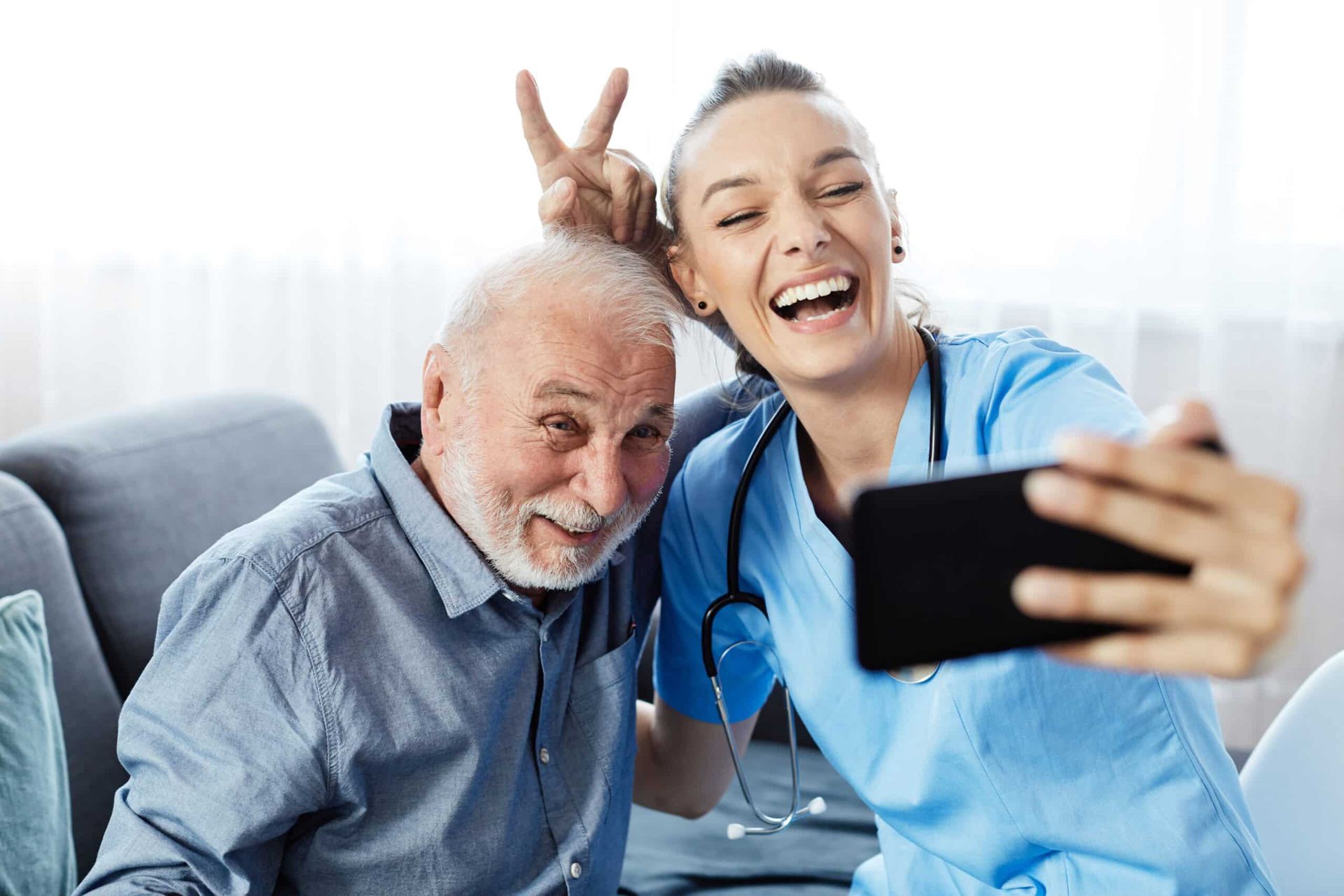 A healthcare worker takes a selfie with an elderly man, both smiling and making the peace sign indoors.