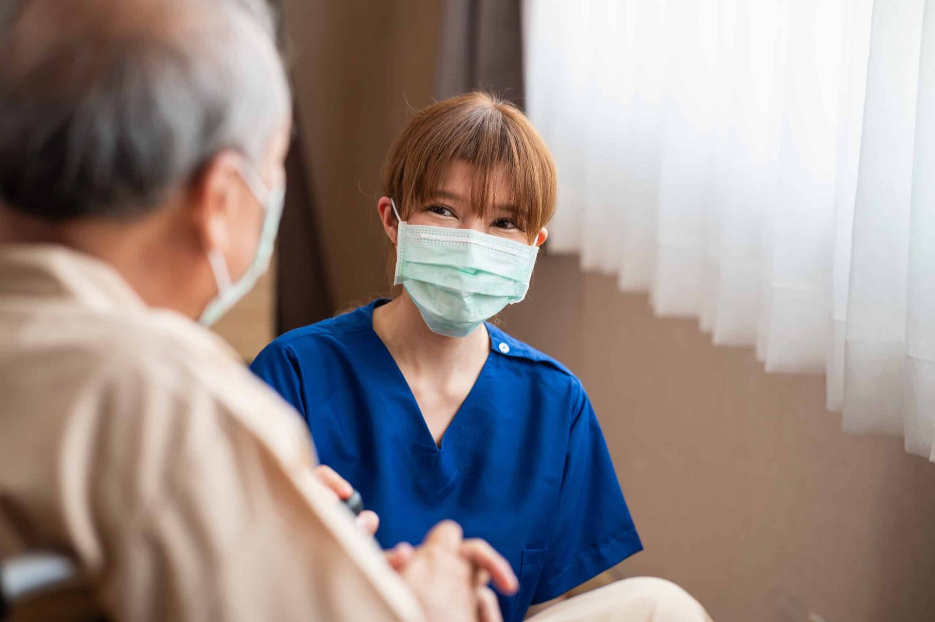 Nurse in blue scrubs and mask smiles at older man in beige shirt indoors.
