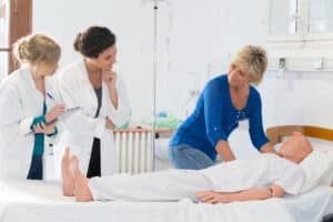 Three medical professionals in white coats observe a patient on a bed in a hospital room.