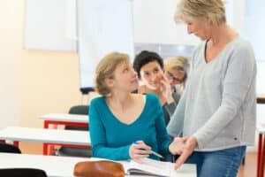 Teacher helping student in a classroom; others look on.
