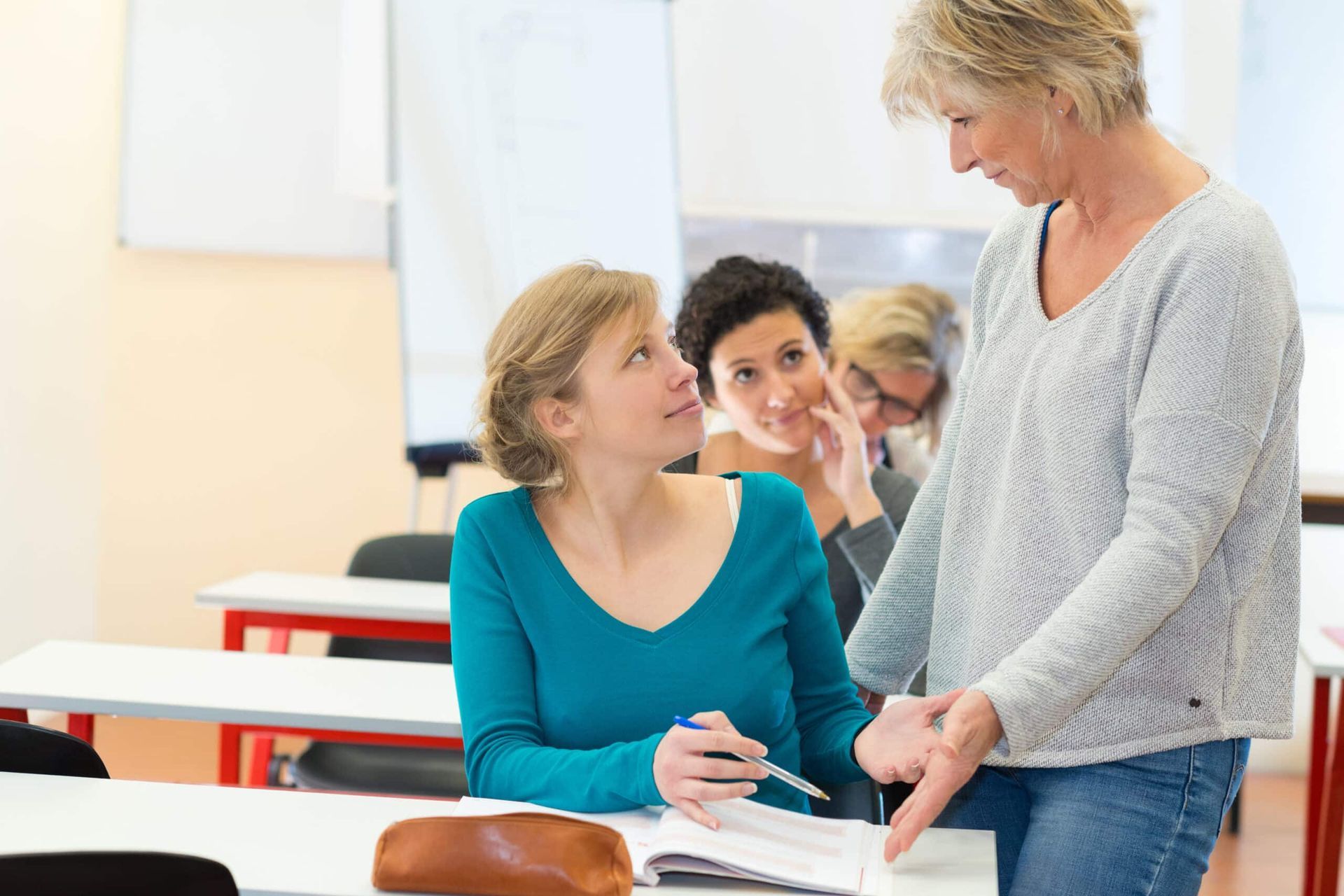 Teacher assisting a student at her desk in a classroom; other students in background.