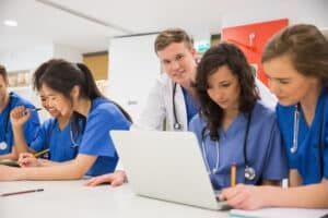 Medical students in blue scrubs gather around a laptop, studying together in a brightly lit room.
