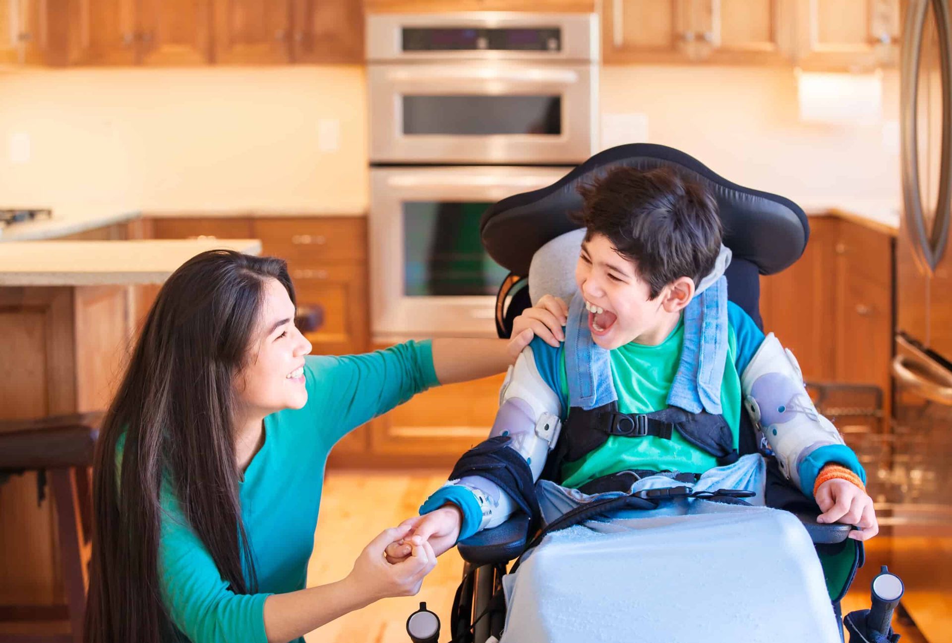 Woman smiles at boy in wheelchair, holding his hand. Kitchen setting, both laughing.