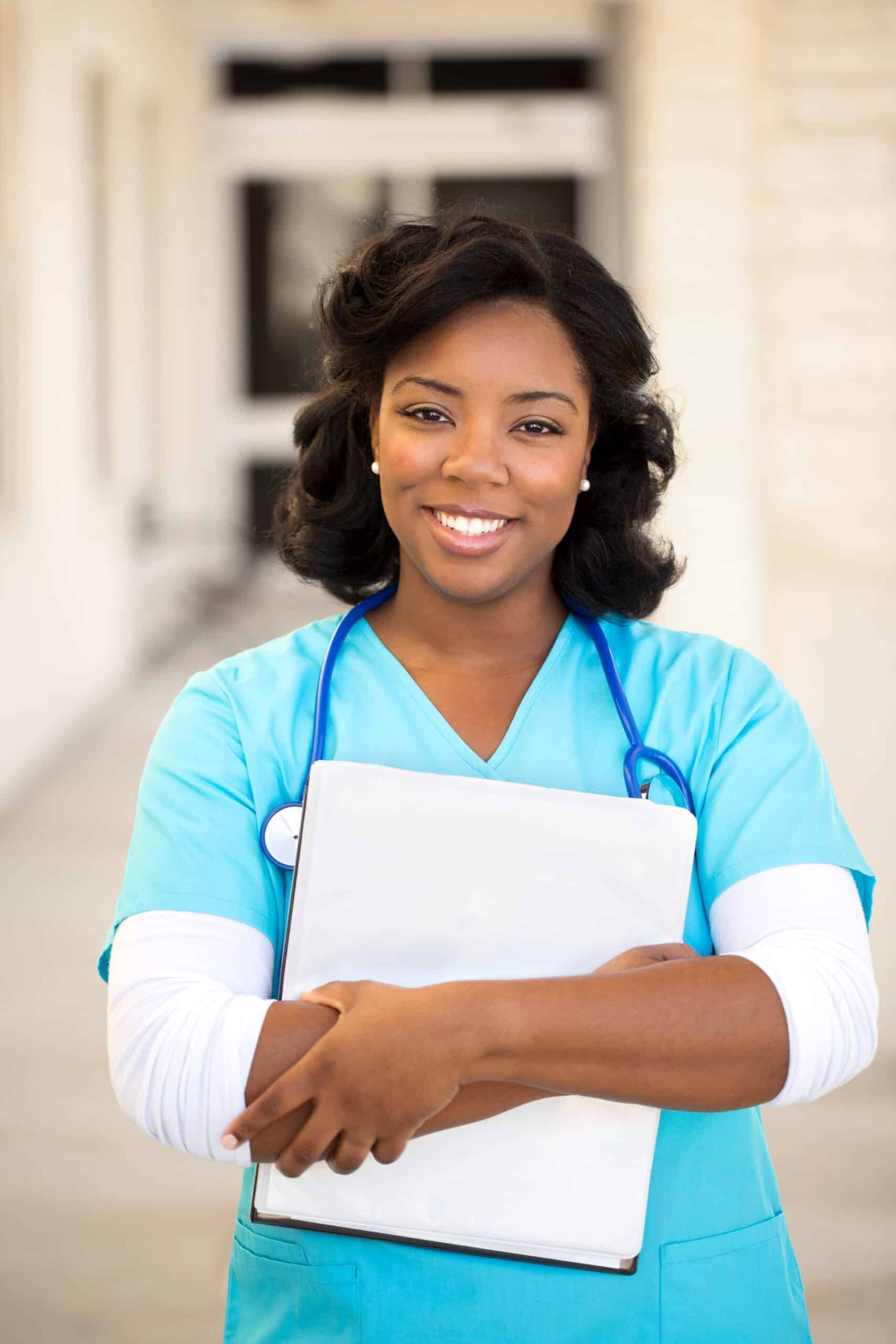 Smiling Black nurse in blue scrubs, holding a clipboard, standing outdoors.