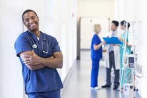 Smiling Black nurse leaning against a wall in a hospital hallway. Medical staff in the background.